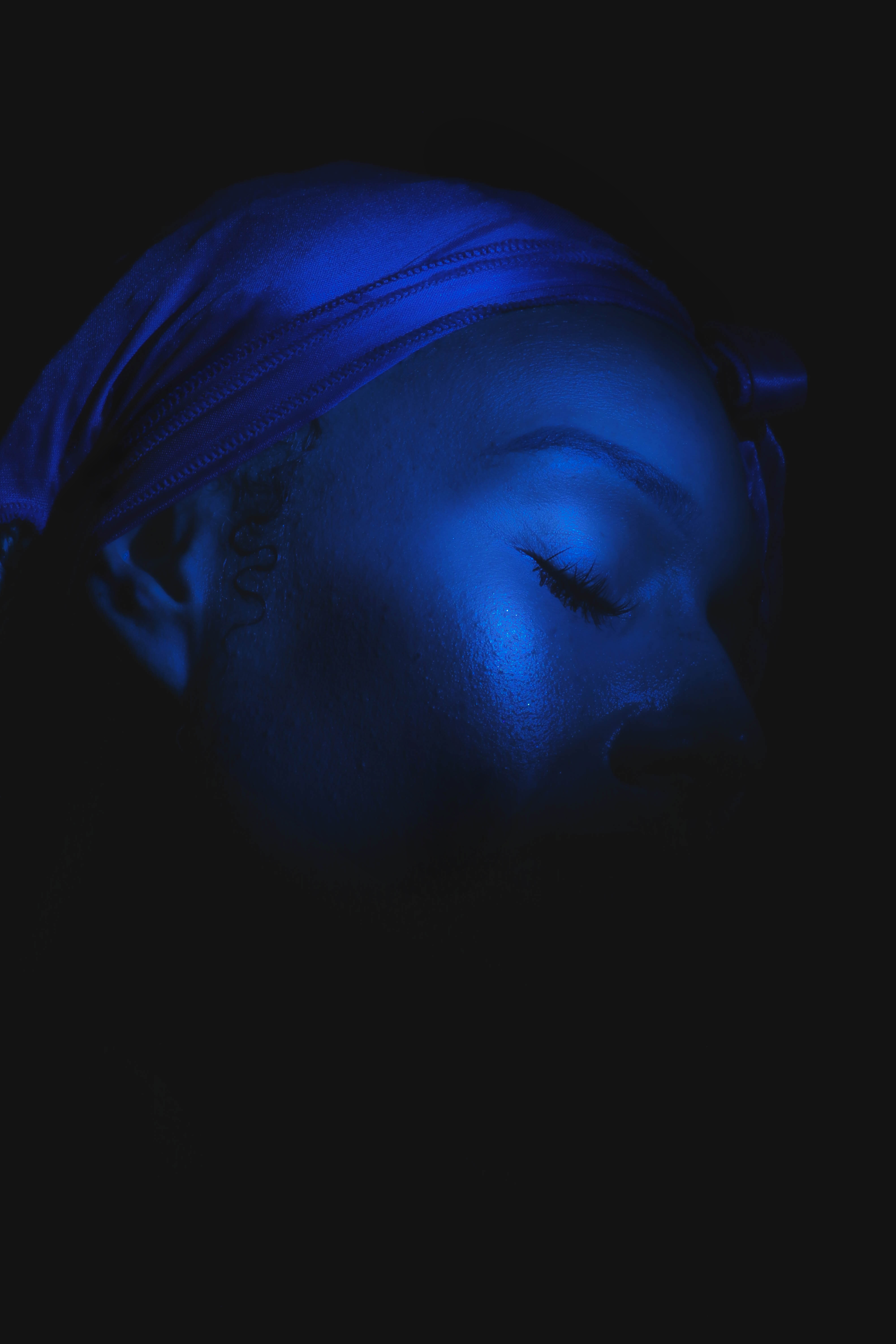 Close-up of a serene Black woman with closed eyes, wearing a blue headscarf against a dark backdrop in a beauty portrait.