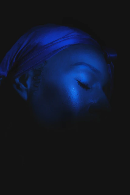 Close-up of a serene Black woman with closed eyes, wearing a blue headscarf against a dark backdrop in a beauty portrait.
