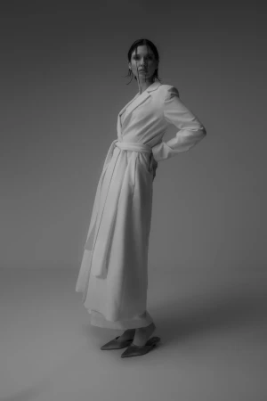 Model in long white belted coat with wet hair standing against grey studio backdrop