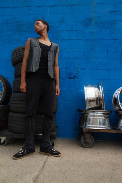 An adult male stands in front of a blue brick wall wearing a black vest, pants, and shoes, with tires and wheels beside him.