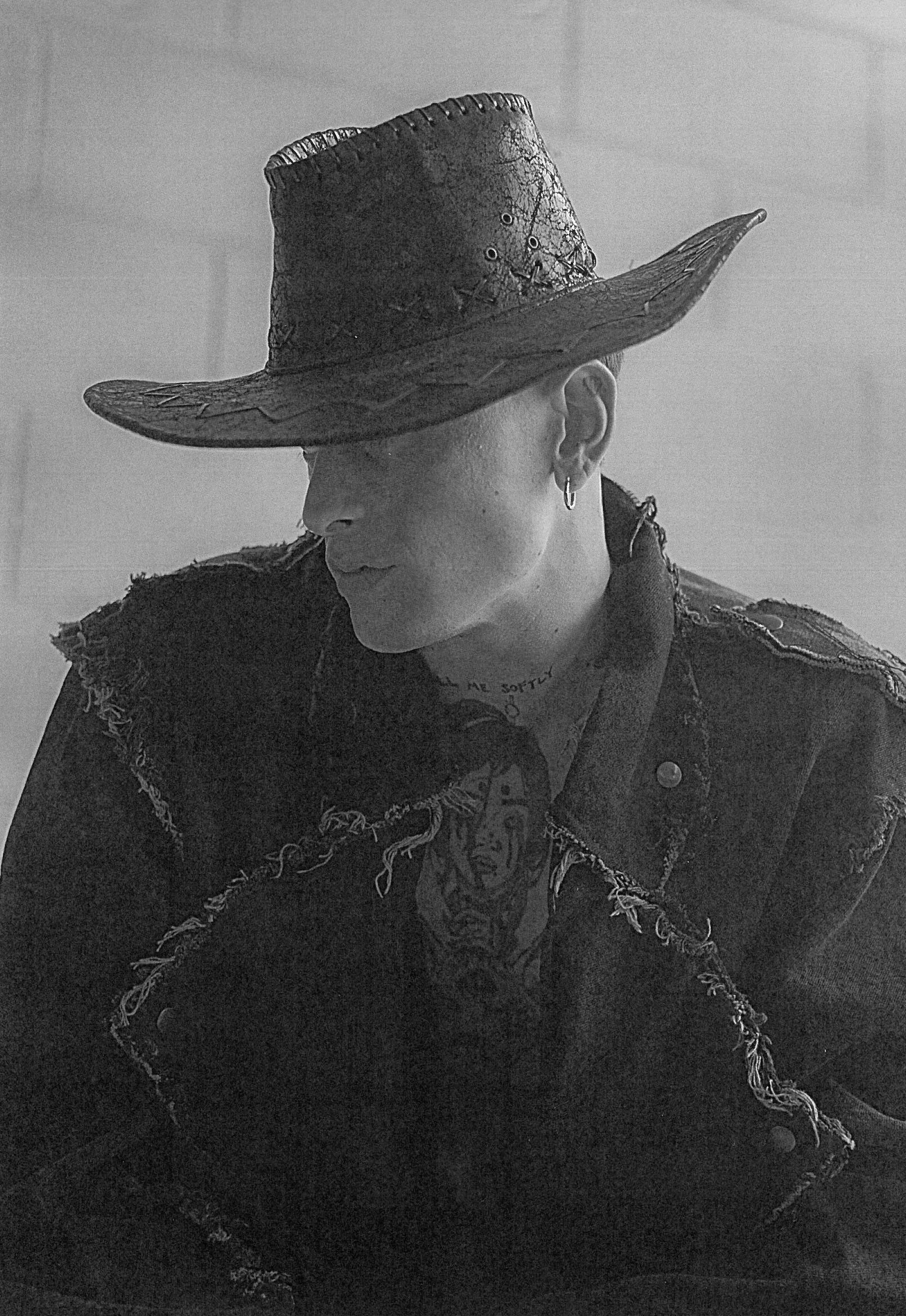 Portrait of a young adult male wearing a cowboy hat and fringed jacket, shot indoors against a pale neutral background in black and white.