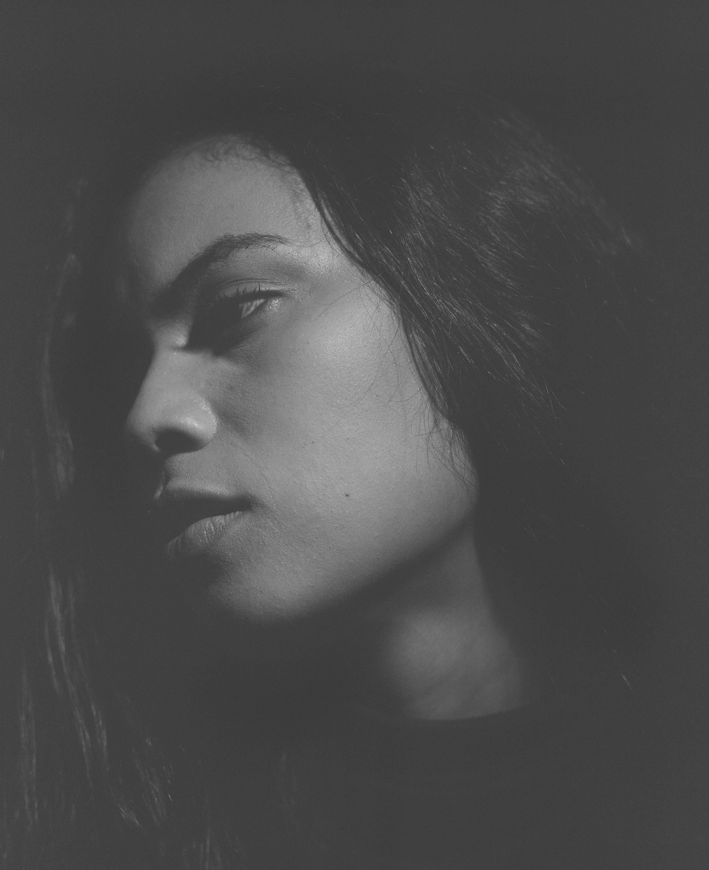 Monochrome portrait of a young adult woman with long hair, glancing slightly down in a dimly lit, indoor environment.