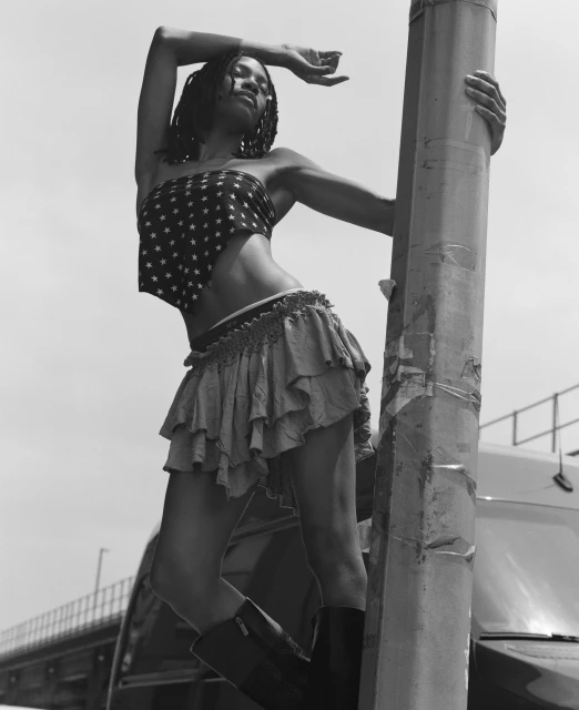 Monochrome portrait of a slender woman in a star-print top and layered skirt, leaning against a pole with a vehicle in the background.