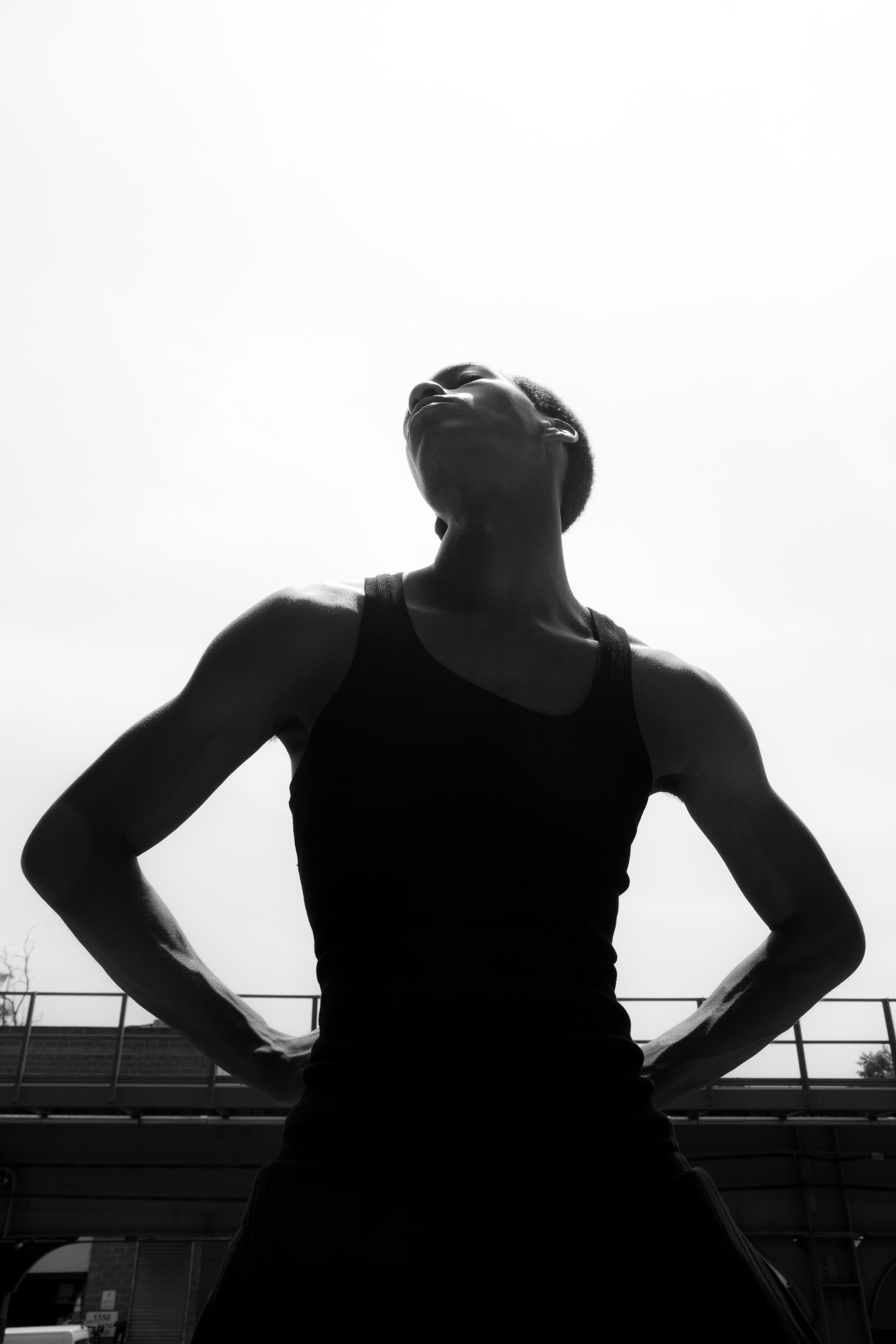 Monochrome shot of a black adult male in tank top with hands on hips looking upward against an out-of-focus background.