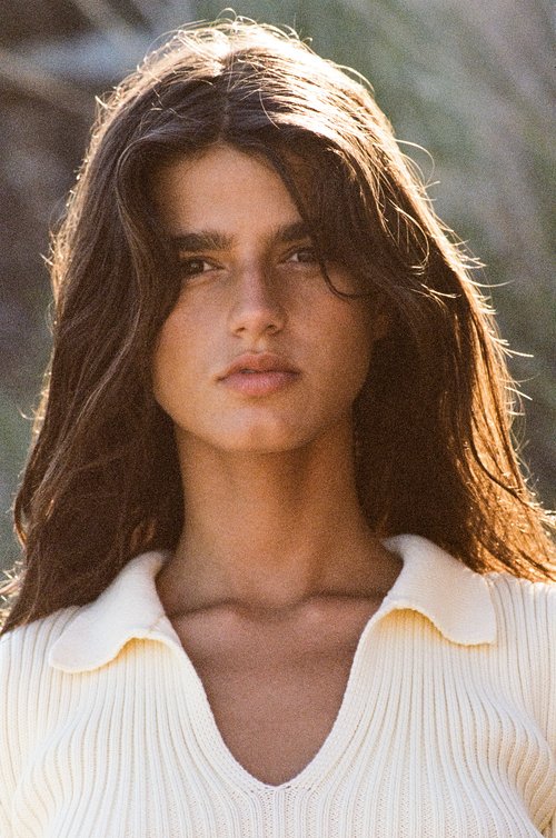 Close-up shot of a young woman with long brown hair wearing a knitted polo shirt, outdoors in soft, natural lighting.