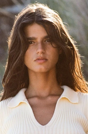 Close-up shot of a young woman with long brown hair wearing a knitted polo shirt, outdoors in soft, natural lighting.