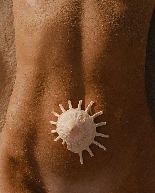 Torso of a person lying on a sand surface, with a seashell placed in the center of the back. Close-up view, warm tones.