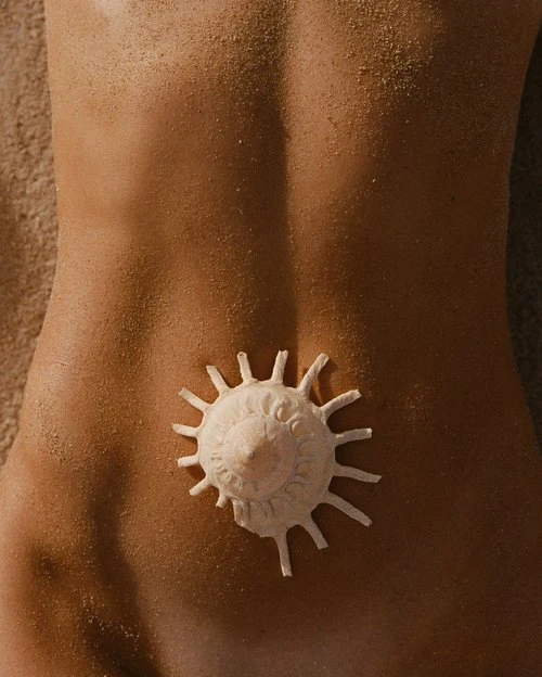 Torso of a person lying on a sand surface, with a seashell placed in the center of the back. Close-up view, warm tones.