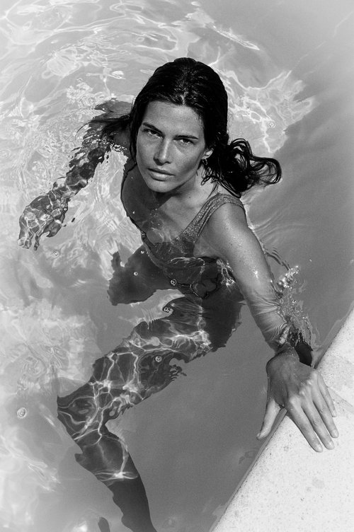 Black and white portrait of a woman in a pool, with the camera angled down, capturing her face, shoulders, and upper body in the water.