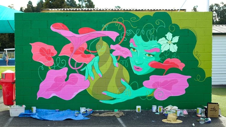 Eye-level outdoor shot of a green brick wall with an illustration of a green woman's face with flowers and leaves. Paint cans are at base.