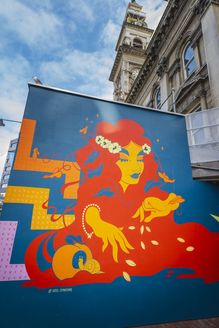 Outdoor mural art of a woman with flowers and a snail on a building in Dunedin, New Zealand, with a historic clock tower in the background.