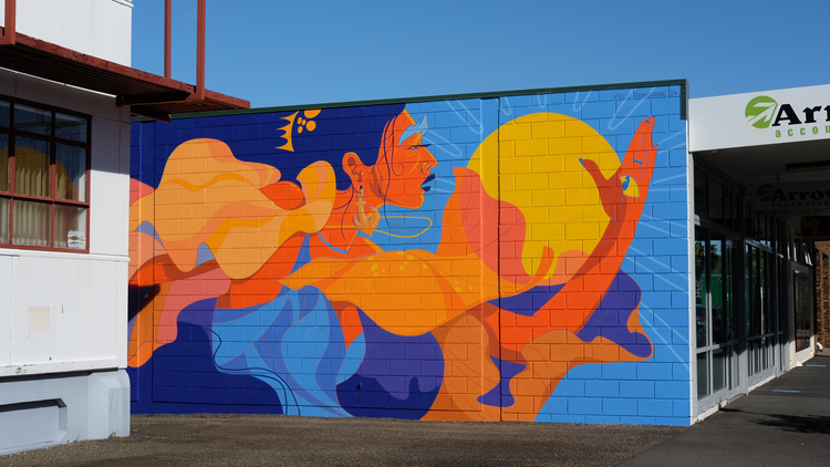 Painted mural featuring an abstract, orange-skinned figure with blue hair and lips holding a yellow sun-like object on a brick wall.
