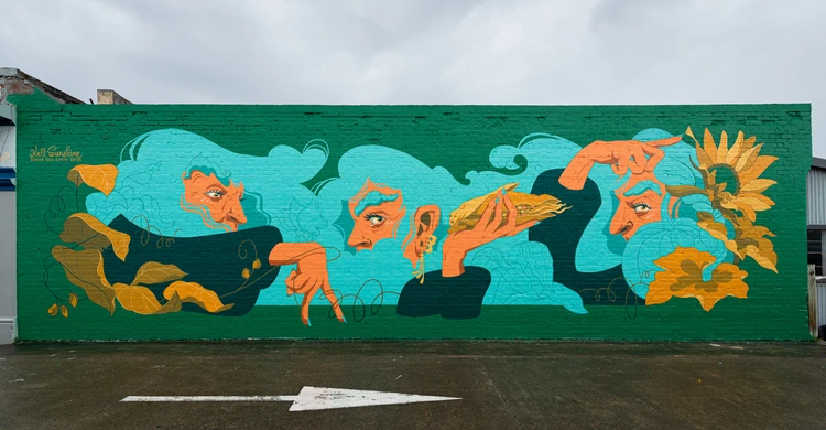 Outdoor mural art on brick wall featuring stylized human faces with flowing hair, hands, and a sunflower in teal, orange, and green hues.
