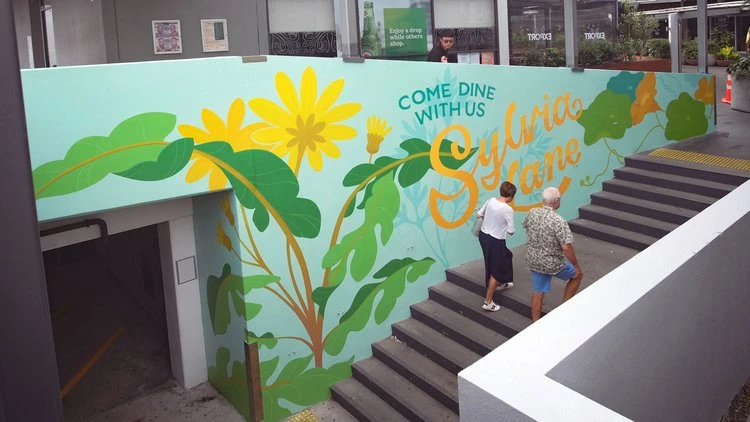Mural illustration by Kelly Spencer on an outdoor wall next to concrete stairs, featuring large yellow flowers and green leaves with typography.