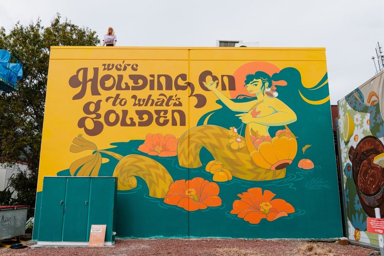 Mural art of woman with flowers in orange and yellow colors with large text. Woman stands on rooftop above the mural art.