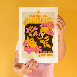 Person holding up a retro-style illustrated art print featuring floral designs and bold text against a yellow wall