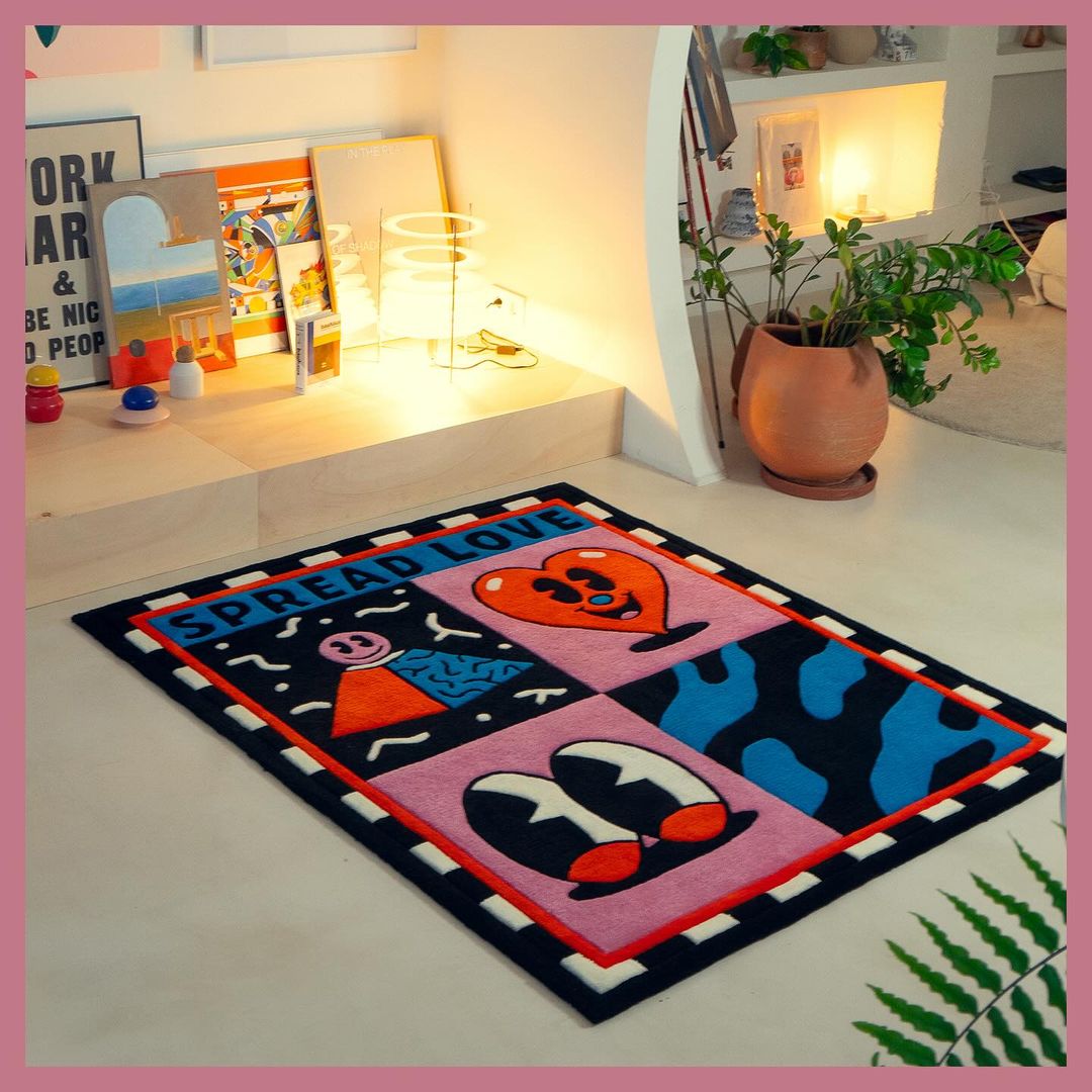 Eye-level shot of a colorful graphic rug with illustrations on a white floor, with a houseplant and framed artwork in the background.