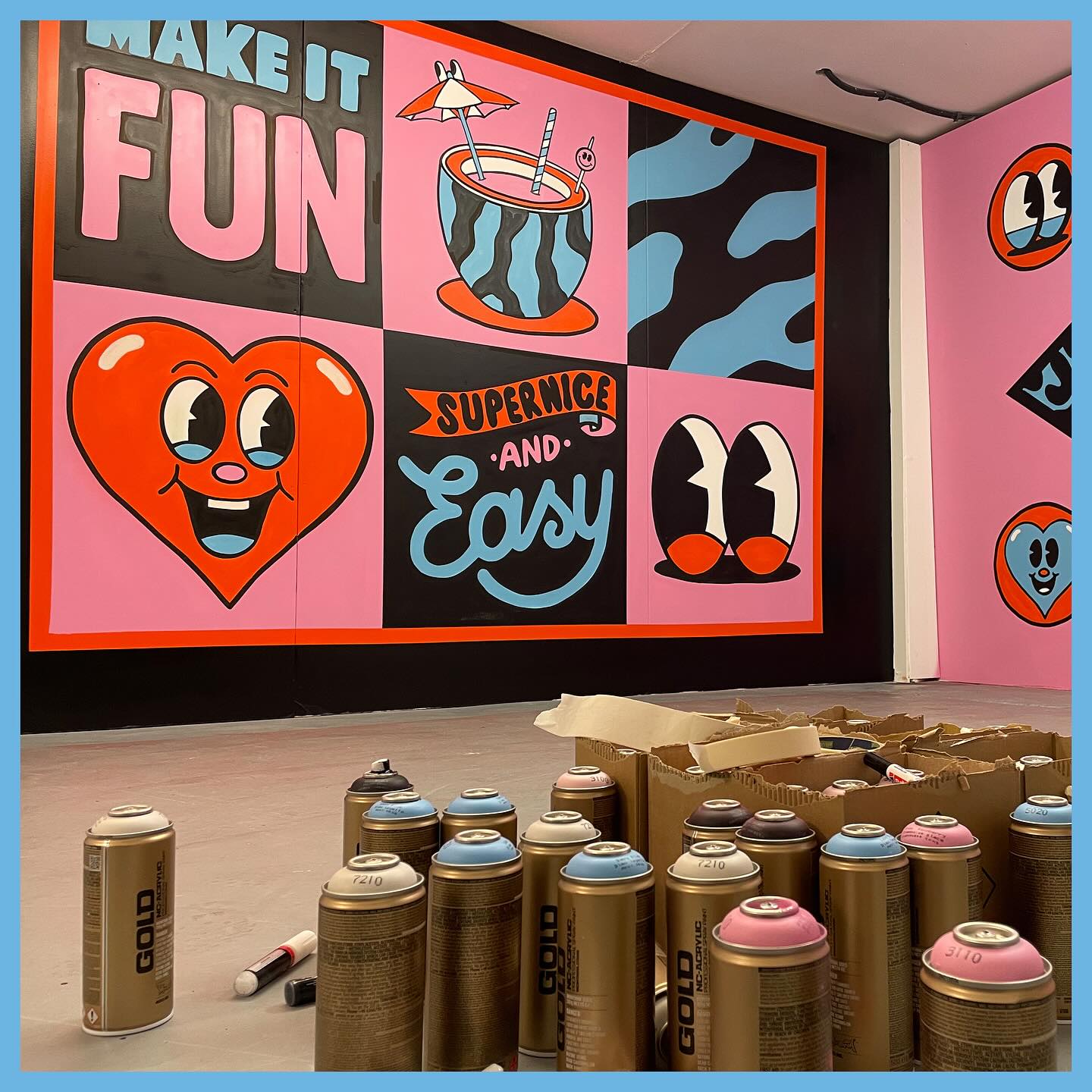 Eye-level shot of a studio wall mural with hand-painted cartoon graphics and text, and spray paint cans in the foreground.