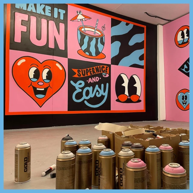 Eye-level shot of a studio wall mural with hand-painted cartoon graphics and text, and spray paint cans in the foreground.