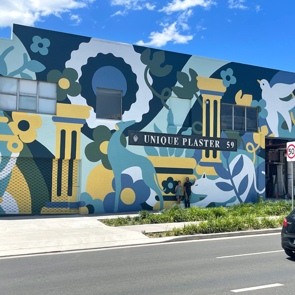 Building exterior featuring a large, colorful mural with stylized animals and plants, observed by two adults, under a bright sky.