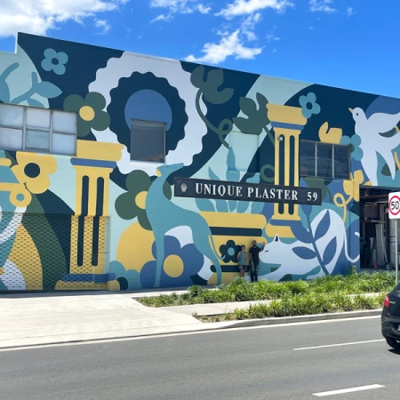 Building exterior featuring a large, colorful mural with stylized animals and plants, observed by two adults, under a bright sky.
