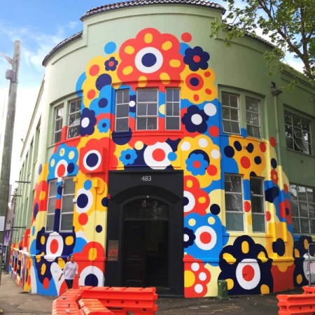 Colorful floral mural on an urban building facade in daylight, featuring red, blue, yellow, and white flower designs with a black doorway.