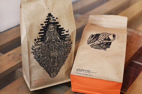 Two brown paper coffee bags with black ink illustrations are displayed against a wood surface in a softly lit, inviting scene.