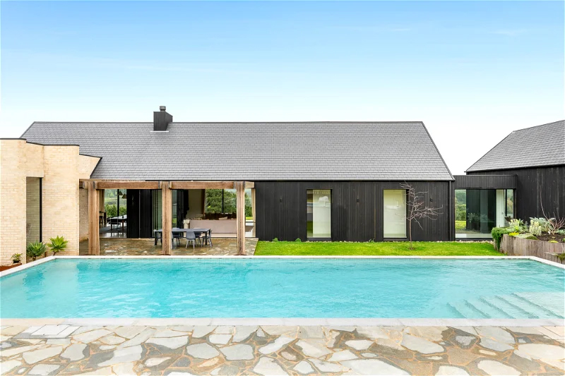 Exterior view of a modern two-story house with a swimming pool and a stone patio under a clear blue sky on a sunny day.