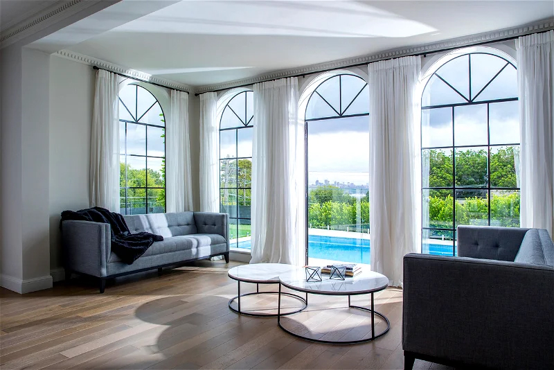 Living room interior with grey sofas, round marble coffee tables, and large arched windows overlooking a pool and landscaped garden.