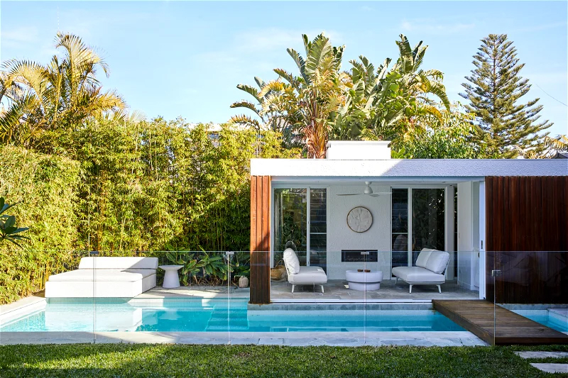 Outdoor living space features a pool, modern building with white furniture, surrounded by lush greenery and bright blue sky.