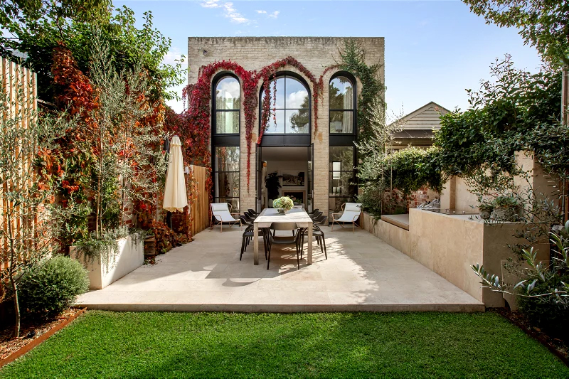 Exterior view of a two-story building with a stone facade, large arched windows, and a courtyard with a dining table and chairs.