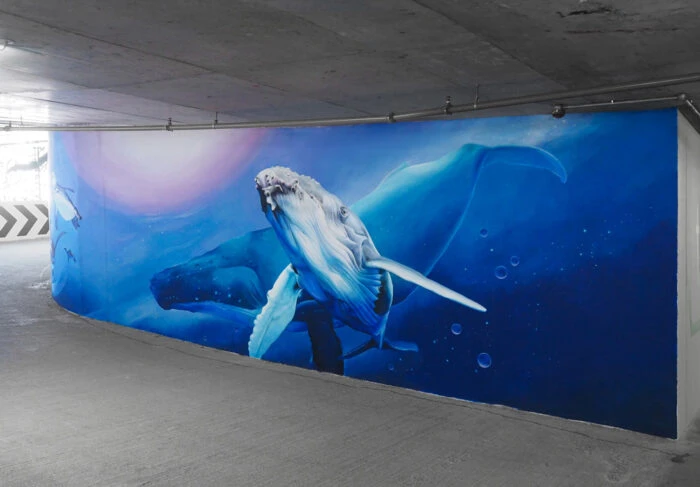 Mural of humpback whale with blue and violet water background on the side wall of a parking structure, shot from an eye-level angle.