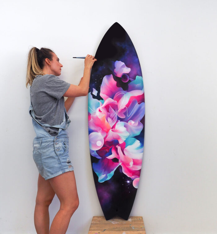 Woman paints a colorful cosmic design on a surfboard in a white photo studio, capturing a blend of art, sport, and fashion.