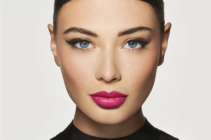 Close up portrait of a woman with blue eyes wearing bold winged eyeliner and bright pink lipstick against a seamless white background.