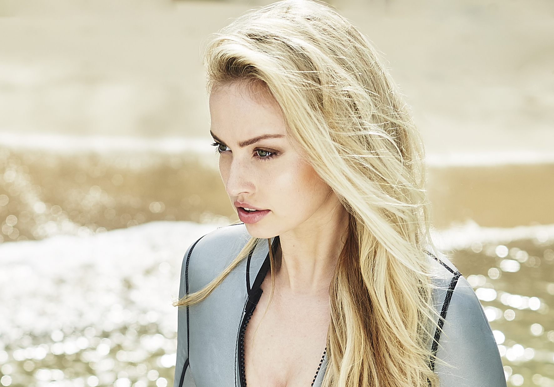 Blonde woman in a wetsuit on a sandy beach with ocean waves, wearing a gray wetsuit in a medium shot, with bright sunlight.
