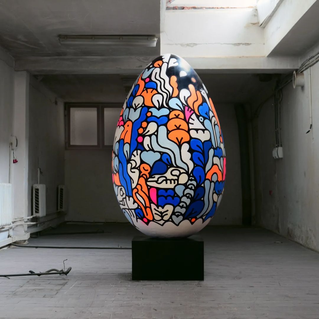 Artwork featuring an Easter egg on a black stand in a spacious room with muted light. Decorative egg is illustrated with patterns and bright colors.