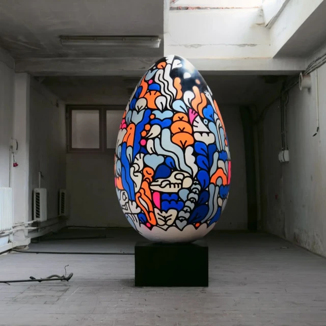 Artwork featuring an Easter egg on a black stand in a spacious room with muted light. Decorative egg is illustrated with patterns and bright colors.