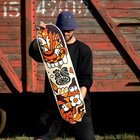 Young man in bucket hat holds skateboard with custom cartoon graphics in front of rusty freight train car outdoors.