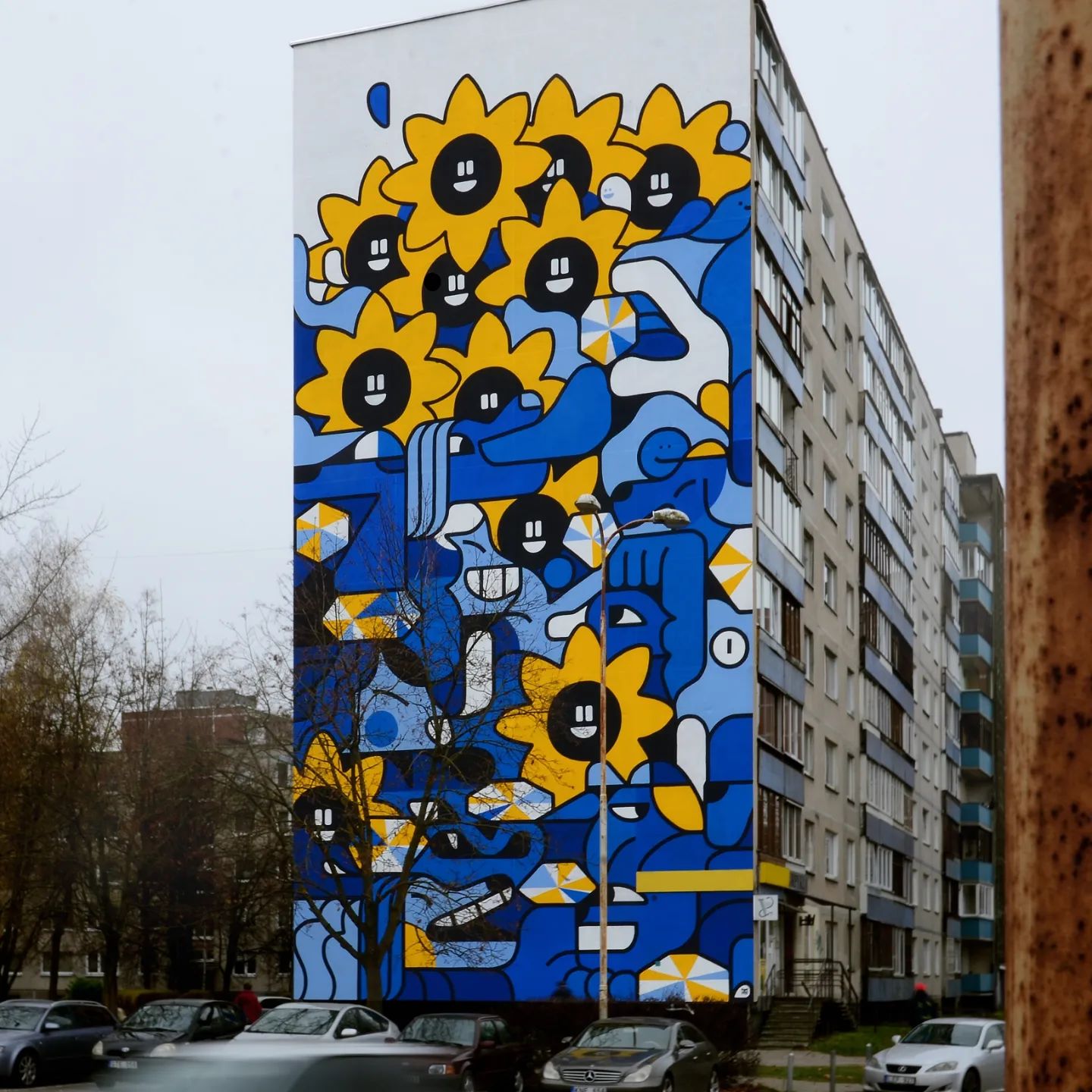 Building facade with mural of sunflowers and smiling faces in shades of blue, yellow, and white; exterior shot.