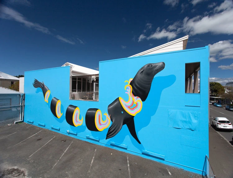 Outdoor mural of a sea lion with colorful, cartoonish cut-out sections on a light blue building under a bright blue sky.