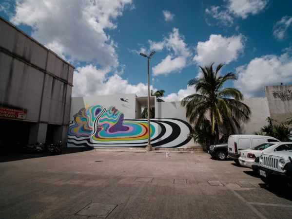 Exterior of a white building with a vibrant mural in a parking area with cars under a partly cloudy blue sky in daylight.