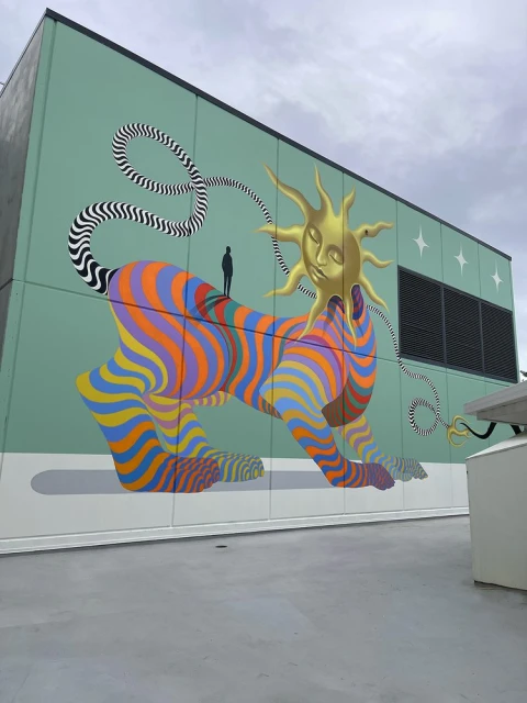Colorful building mural of striped quadruped with golden sun head & tiny figure standing on back under cloudy sky.