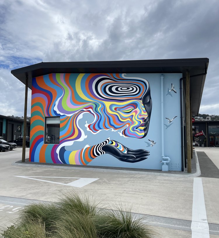 Outdoor mural featuring a profile of a woman's face in shades of black and gray, with colorful waves flowing into a cupped hand.
