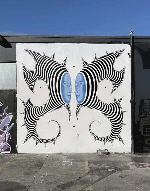Outdoor mural art features two blue-faced figures with striped headpieces facing each other, set against a light gray wall and blue sky.