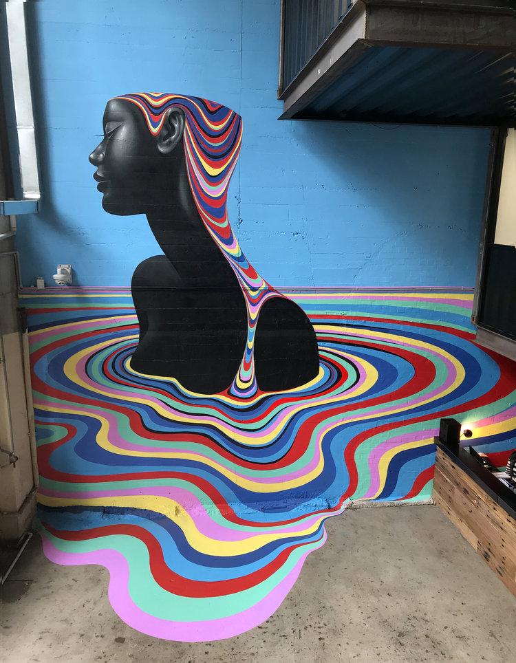 Mural of a black female figure with rainbow-colored hair and water ripples, creating a captivating artwork on a light blue wall indoors.