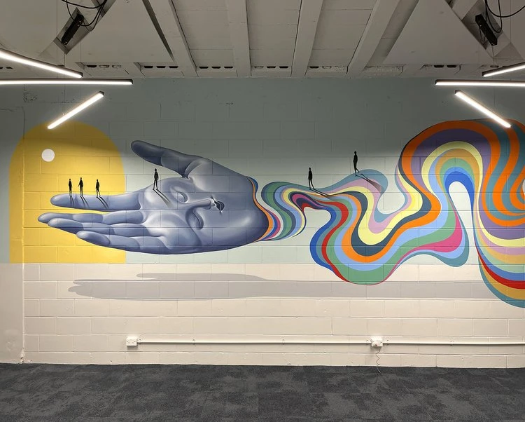 Art wall featuring a large hand mural with tiny figures walking along a colorful, wavy path. Interior with visible lighting.