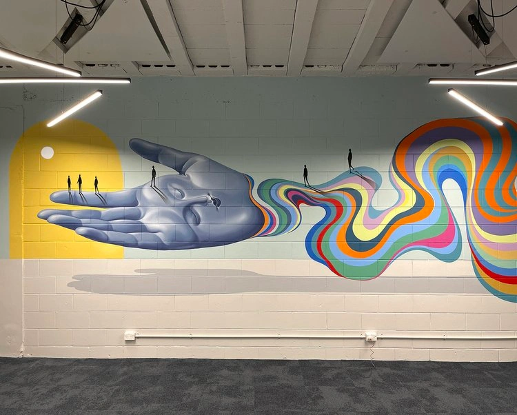 A large wall mural featuring a surreal giant hand emerging from colourful wavy lines on a painted pale blue background.