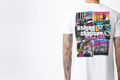 Man wearing a white t-shirt with a colorful graphic illustration on the back and tattoos on his arms against a plain white background.