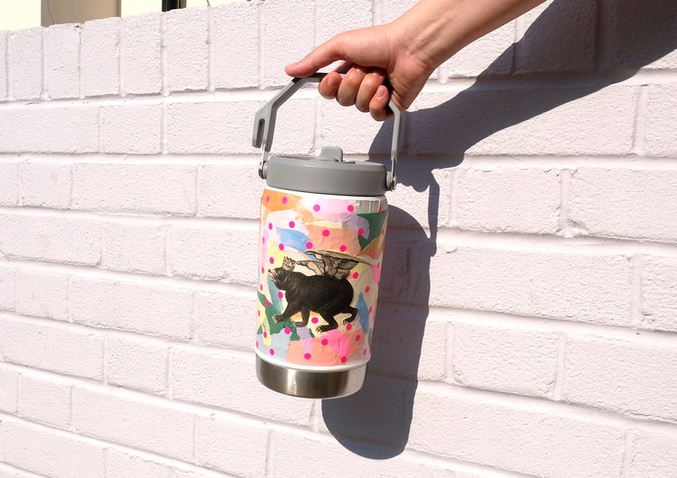 Person holding a decorated stainless steel water bottle with a whimsical winged bear design against a bright white brick wall.