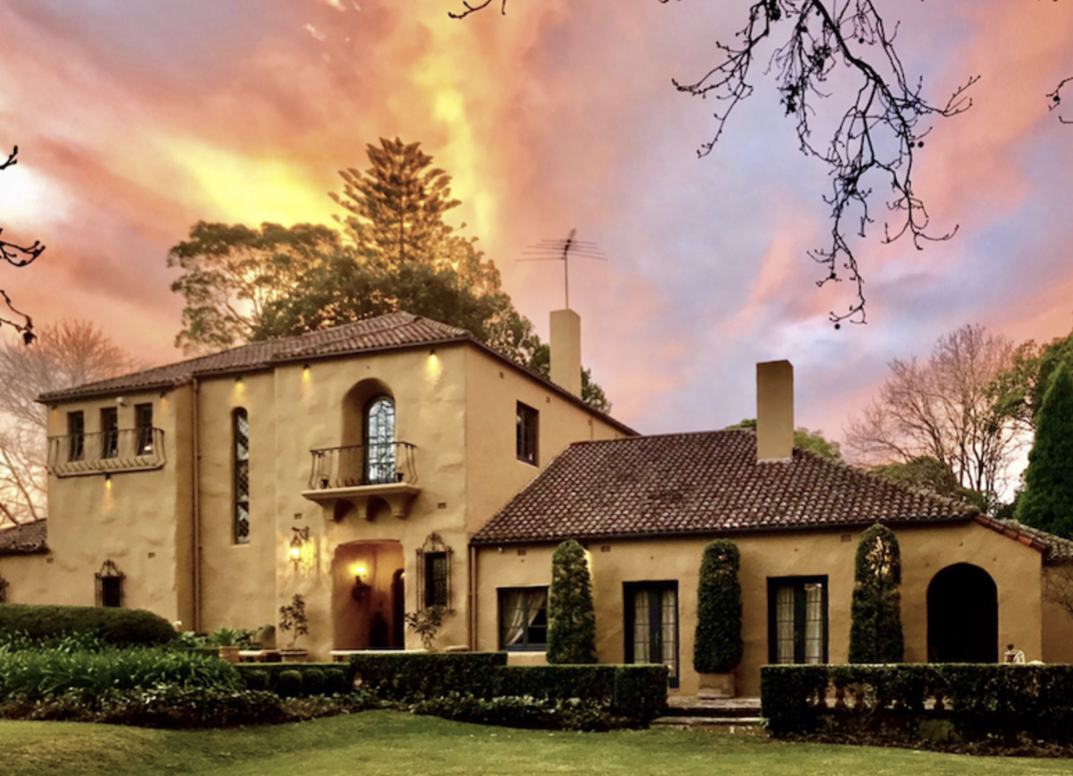 Exterior of a tan stucco Mediterranean-style house with a red tile roof, green lawn, and shrubbery under a pink and orange sunset sky.